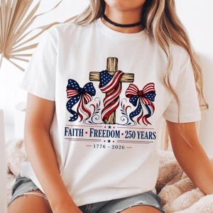 Op de afbeelding: Een wit T-shirt met korte mouwen en een afbeelding van een houten kruis gedrapeerd met een lint in de kleuren van de Amerikaanse vlag, geflankeerd door twee patriottische strikken. Daaronder staat de tekst FAITH • FREEDOM • 250 YEARS en 1776 - 2026.