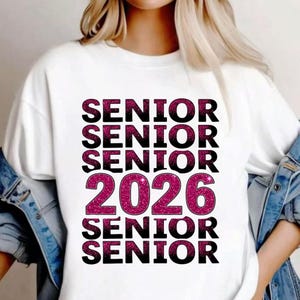 Op de afbeelding: Witte sweatshirt met het woord "SENIOR" meerdere keren herhaald in een gestapeld ontwerp. Het jaar "2026" staat in het midden, met een roze glittereffect. De sweatshirt heeft een casual stijl.