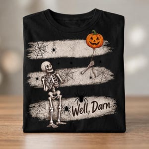 Könnte beinhalten: Ein schwarzes T-Shirt mit einer Grafik eines Skeletts, das eine Schnur hält, an der ein orangefarbener Kürbisballon befestigt ist. Das Design zeigt drei horizontale, abgenutzte weiße Streifen, Spinnennetze und Spinnen sowie den Text Well, Darn...