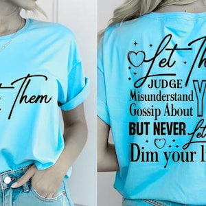 Puede incluir: Camiseta azul claro con la frase "Let Them" en fuente negra en la parte delantera. La parte trasera de la camiseta tiene el texto "Let Them" con las palabras "JUDGE, Misunderstand, Gossip About, BUT NEVER, Dim your light!"