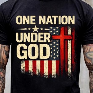Könnte beinhalten: Schwarzes T-Shirt mit dem weißen Schriftzug "ONE NATION UNDER GOD", einem amerikanischen Flaggen-Design und einem roten Kreuz. Das Design hat einen Used-Look.