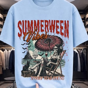 Peut inclure: T-shirt bleu clair avec un motif squelette sur une chaise longue sous un parasol rouge, tenant un verre de vin. Le texte dit "SUMMERWEEN Vibes" et "SPOOKY YEAR ROUND". Un chat noir est assis sur un coffre.