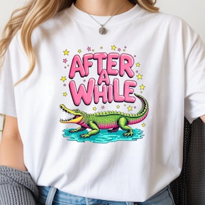 Peut inclure: T-shirt blanc avec un alligator de dessin animé vert et rose, et les mots "AFTER A WHILE" en lettres roses à bulles. Le motif comprend des étoiles jaunes et un effet d'eau bleu.