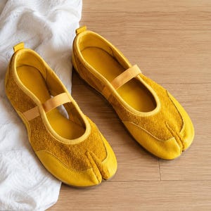 May include: A pair of yellow split-toe shoes with a strap across the top. The shoes have a suede-like texture on the toe and a smooth leather-like material on the rest of the shoe. The shoes are on a wooden surface.