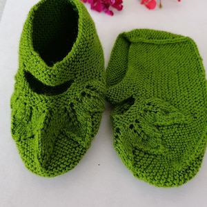 Peut inclure: Une paire de chaussons tricotés à la main d'un vert vif, présentant un motif texturé au point mousse et un design décoratif en forme de feuille sur le devant. Les chaussons ont un bout arrondi, une ouverture basse et une structure souple.