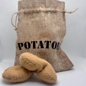 May include: A hessian sack printed with the word "POTATO" in black, alongside two tan, stuffed potato-shaped toys. The toys feature visible stitching, and the sack has a drawstring closure.