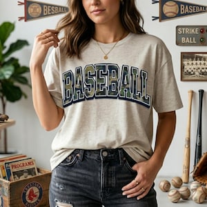 May include: A light grey t-shirt featuring the word "BASEBALL" in large, camouflage-style lettering. The shirt has a crew neck and short sleeves. The model is wearing dark-wash jeans. Baseball-themed decor is visible in the background.