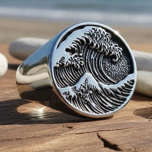 May include: A silver signet ring with a detailed wave design. The ring is round with a polished finish, and the wave is depicted in a circular emblem. The ring is displayed on a wooden surface with pebbles and driftwood.