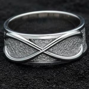 Könnte beinhalten: Ein silberner Ring mit einem Unendlichkeitszeichen. Der Ring hat ein strukturiertes Band mit polierten Kanten. Das Unendlichkeitszeichen ist ein wichtiges Designelement, das einen Hauch von Eleganz verleiht. Der Ring wird auf einer dunklen, strukturierten Oberfläche präsentiert.