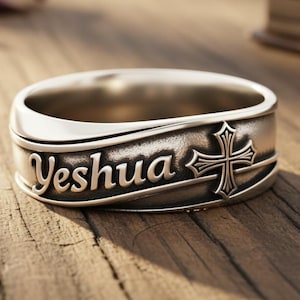 May include: Silver-toned ring with the word "Yeshua" and a cross design. The ring is on a wooden surface with a blurred background that includes a book and flower petals. The ring is a religious item.
