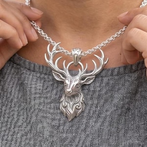 Puede incluir: Un collar de tono plateado con un colgante detallado de una cabeza de ciervo con grandes astas ramificadas. El colgante cuelga de una cadena gruesa y texturizada de tono plateado que descansa sobre una prenda de tela gris. El metal es pulido.