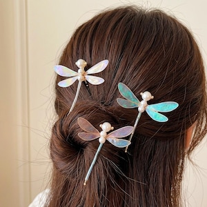 May include: Three dragonfly hair pins featuring iridescent, translucent wings and white pearl-like beads for the head and thorax. Each pin has a slender metal stem, with the wings displaying shifting hues of blue, pink, and purple light.