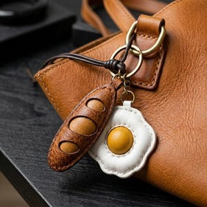 May include: A brown leather keyring featuring two charms: a textured brown baguette-shaped charm and a white and yellow fried egg-shaped charm. The keyring is attached to a brown leather bag with a gold-toned metal ring and a dark brown cord.