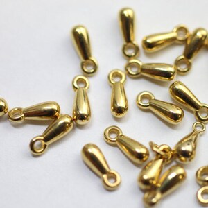 Könnte beinhalten: Eine Ansammlung kleiner, glänzend goldfarbener Metallanhänger in Tropfenform. Jeder Anhänger hat eine glatte, reflektierende Oberfläche und eine kreisförmige Öse an der Oberseite zur Befestigung, verstreut auf einer hellen Oberfläche.