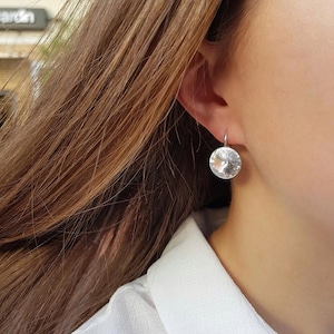 Könnte beinhalten: Nahaufnahme eines silbernen Ohrrings mit einem großen, runden, klaren Kristall. Der Kristall ist facettiert und reflektiert das Licht. Der Ohrring ist an einem silbernen Haken befestigt. Der Hintergrund ist verschwommen, mit Haaren und einem weißen Hemd.