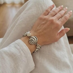 Könnte beinhalten: Ein silberfarbenes Gliederarmband mit sich wiederholenden kreisförmigen Spiralmotiven, die durch kleine ovale Metallsegmente verbunden sind. Das Armband hat eine matte, gebürstete Oberfläche und liegt auf einem Handgelenk vor weißem Stoff.