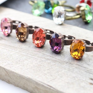 Könnte beinhalten: Eine Sammlung verstellbarer Ringe mit großen, oval geschliffenen Facettenkristallen in verschiedenen Farben wie Rosa, Bernstein, Orange, Violett und Gold. Jeder Kristall ist in einer bronzefarbenen Metallfassung mit schmaler Ringschiene.