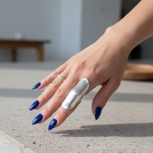 Könnte beinhalten: Ein großer, glatter, silberfarbener Metallpanzerring, der den Mittelfinger bedeckt und ein geschwungenes, längliches Design aufweist. Ein dünner, texturierter goldfarbener Bandring wird am Ringfinger getragen. Die Fingernägel sind dunkelblau lackiert.