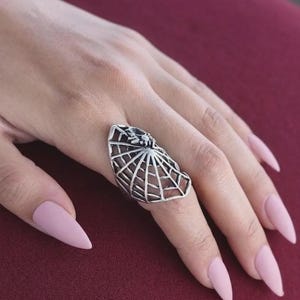 Könnte beinhalten: Ein silberfarbener Ring mit einem durchbrochenen Spinnennetz-Design, das sich über den Finger erstreckt. Die Metalloberfläche weist in den vertieften Bereichen ein dunkles, oxidiertes Finish auf, das das komplexe Netzmuster und eine kleine Spinne hervorhebt.