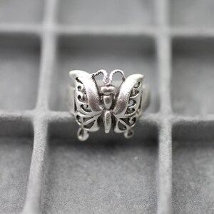 Könnte beinhalten: Ein silberfarbener Metallring mit einem Schmetterlingsmotiv, das filigrane Ausschnitte an den Flügeln und zwei kleine Fühler an der Oberseite aufweist. Das Schmetterlingsdesign ist auf einer glatten, festen Ringschiene zentriert.