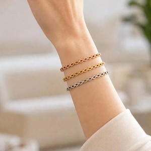 Könnte beinhalten: Drei dünne Armbänder, die zusammen getragen werden und jeweils ein sich wiederholendes Unendlichkeits- oder Ovalmuster aufweisen. Das Set besteht aus einem roségoldfarbenen, einem goldfarbenen und einem silberfarbenen Armband am Unterarm.