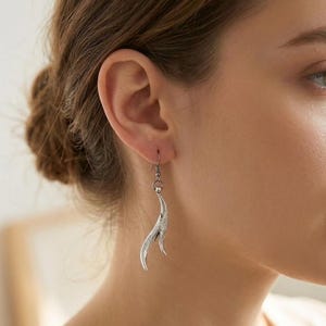 Könnte beinhalten: Ein silberfarbener Hängeohrring mit einer geschwungenen, texturierten, flammenartigen Metallform, die an einem einfachen Haken befestigt ist. Der Ohrring hat ein mattes, leicht oxidiertes Finish, das die feinen Oberflächendetails des Designs betont.