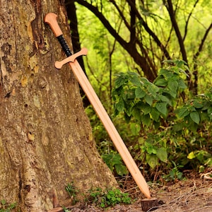 May include: A wooden sword with a light brown blade and crossguard, featuring a black textured grip and a rounded pommel. The sword is positioned diagonally against the rough, textured bark of a large tree trunk in a wooded outdoor setting.