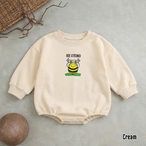 May include: Cream-colored baby romper with long sleeves. The romper features a graphic of a cartoon bee lifting weights, with the text "BEE STRONG!". The garment is made of a soft material.