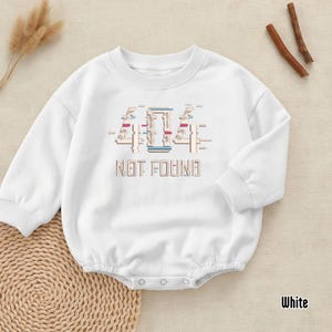 Könnte beinhalten: Weißer Baby-Strampler mit langen Ärmeln und rundem Halsausschnitt. Auf der Vorderseite steht der Text "404 NOT FOUND" in einem pixeligen Design mit blauen, roten und beigen Akzenten. Der Strampler hat Druckknöpfe am unteren Ende.