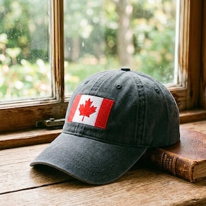 May include: A charcoal gray baseball cap featuring the Canadian flag. The flag is a red maple leaf on a white background, set within a red border. The hat is resting on a wooden surface near a window.