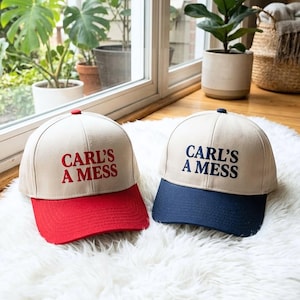 Puede incluir: Dos gorras de béisbol con el texto "CARL'S A MESS". Una gorra tiene la visera roja y el texto rojo, la otra tiene la visera azul marino y el texto azul marino. Ambas gorras son beige y están sobre una superficie blanca y esponjosa.