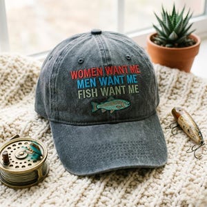 May include: A dark grey baseball cap with the embroidered text "WOMEN WANT ME, MEN WANT ME, FISH WANT ME." A teal and blue fish is embroidered below the text. The cap is displayed on a cream-coloured knitted blanket, with fishing tackle in the background.