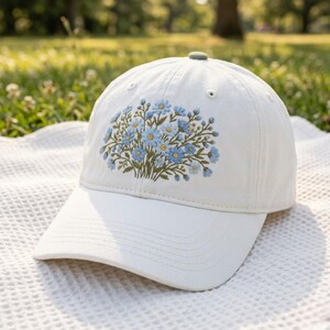 Puede incluir: Gorra de béisbol blanca con visera curva. Presenta un diseño bordado de flores azules y amarillas con tallos verdes. Tiene un botón verde en la parte superior y está sobre una manta blanca.