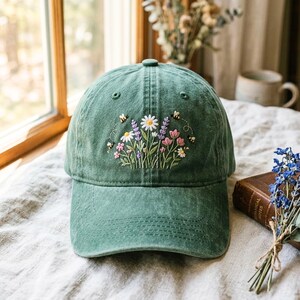 Peut inclure: Casquette de baseball verte avec un motif floral brodé de marguerites, de lavande et de fleurs roses, avec de petites abeilles brodées bourdonnant autour des fleurs. La casquette est en tissu doux et usé.