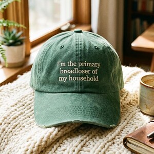 May include: A green baseball cap with the embroidered text "I'm the primary breadloser of my household." The cap is on a cream knitted blanket. The cap has a curved peak and a classic six-panel design. The fabric has a distressed look.