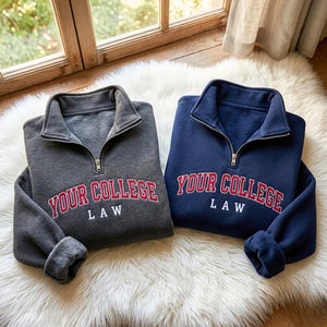 Könnte beinhalten: Zwei Sweatshirts mit Viertelreißverschlusskragen werden auf einer weißen, flauschigen Oberfläche gezeigt. Eines ist dunkelgrau, das andere marineblau. Beide haben den Text "YOUR COLLEGE LAW" in roten Blockbuchstaben.