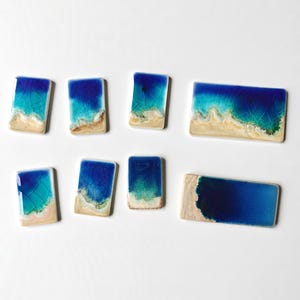 May include: A collection of rectangular ceramic pieces with a beach scene design. The pieces feature a gradient of blue and turquoise representing the ocean, with a tan and white sandy beach at the bottom.
