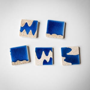 Puede incluir: Cinco posavasos cuadrados de cerámica con un esmalte azul y un borde ondulado color arena. Los posavasos tienen un acabado brillante y están dispuestos sobre un fondo blanco. El esmalte azul varía en patrón.