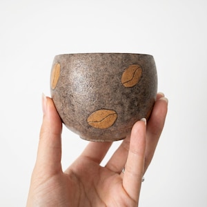 Op de afbeelding: Een kleine, ronde, bruine keramische kom met een getextureerd oppervlak. De kom is versierd met verschillende grote, bruine koffieboonontwerpen. De kom wordt in de hand van een persoon tegen een witte achtergrond gehouden. De kom wordt waarschijnlijk gebruikt om koffie of thee uit te drinken.