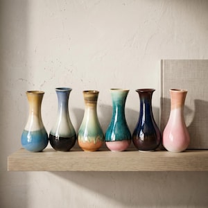 May include: A set of six small ceramic bud vases with a flared rim and a rounded base. Each vase features a unique reactive glaze finish in varying color combinations including blue, brown, teal, pink, and cream, arranged in a single row.