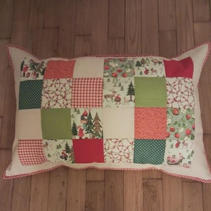 Op de afbeelding: Rechthoekig patchwork kussen met een witte achtergrond en een rood-witte ruitrand. Het patchwork ontwerp heeft vierkanten van rode, groene en witte stof met kerstthema patronen, waaronder bomen, herten en bessen.