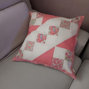 Puede incluir: Un cojín cuadrado con un diseño de patchwork que combina cuadrados de tela blanca lisa con cuadrados estampados en tonos coral, crema y verde, incluyendo pequeños motivos de pájaros azules y detalles de tela con lunares blancos.