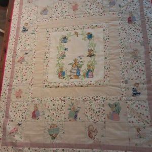 Könnte beinhalten: Cremefarbene Steppdecke mit einem zentralen Quadrat mit Peter Hase und anderen Figuren. Die Steppdecke hat ein Blumen- und Hasenmuster mit einem rosa Rand und roter Einfassung. Für ein Baby oder Kind.