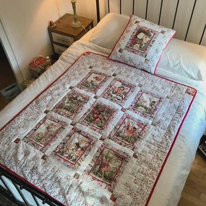 May include: A white quilt and matching pillow set with a floral and fairy design. The quilt features framed images of fairies and flowers, bordered in red. The bedspread is on a black metal bed frame.