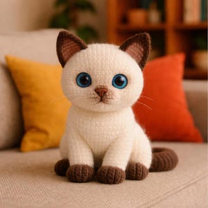 以下が含まれることがあります： 手作りのかぎ針編みシャム猫のぬいぐるみ。猫はクリーム色の体、茶色の耳、足、尾、そして印象的な青い目をしています。ベージュのソファに座っており、背景にはカラフルなクッションがあります。