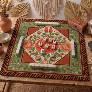 May include: A Mahjong game set on a decorative mat with a floral design. The mat features a central red flower surrounded by green and orange patterns. The set includes tiles and is displayed on a wooden table, with a wooden box and decorative items in the background.