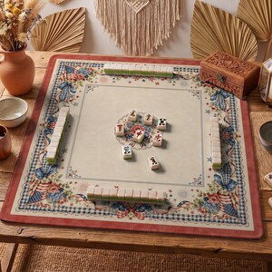 Könnte beinhalten: Eine quadratische Mahjong-Matte mit cremefarbenem Zentrum und einem Rand, der mit rot-weiß-blauen Blumenmustern, karierten Bändern und amerikanischen Flaggenmotiven verziert ist. Auf der Matte sind mehrere weiße Mahjong-Spielsteine angeordnet.