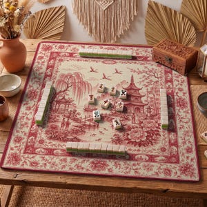 May include: A Mahjong game set on a red and cream-colored mat with a floral and Asian-inspired landscape design. The set includes tiles and is arranged on a wooden table, with a decorative wooden box in the background.
