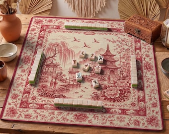 May include: A Mahjong game set on a red and cream-colored mat with a floral and Asian-inspired landscape design. The set includes tiles and is arranged on a wooden table, with a decorative wooden box in the background.