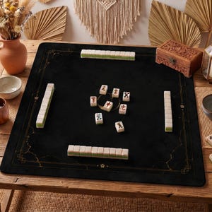 May include: A square black mahjong mat featuring gold-colored decorative border lines and floral accents. The mat is arranged with four rows of white and green mahjong tiles along the edges and several individual tiles placed in the center.